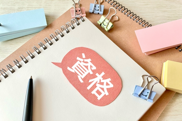 社内ニートの資格勉強を解説する記事のアイキャッチ｜リングノートに赤い吹き出しで白抜きの「資格」の文字。付箋やクリップ、ペンが並ぶ木目の机の上。暇な時間を資格取得の準備に充てる様子を表現