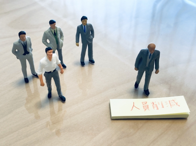 早期退職その後の50代のリアルのアイキャッチ｜「人員削減」のメモの前で、スーツ姿のミニチュア人形の集団から一人だけ離れてうつむく男性。会社を去る50代の孤独と決断を象徴する構図