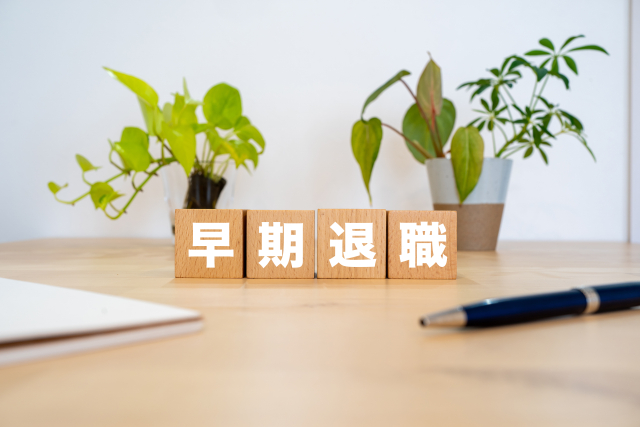 57歳の早期退職「末路」がテーマの記事のアイキャッチ｜観葉植物とノートに囲まれたデスクに「早期退職」と書かれた木製ブロックが並ぶ様子。定年を目前にした決断のタイミングと、静かに人生の岐路と向き合う心境を表現